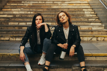 Portrait of two girls models in stylish casual clothes sitting on stairs with bottle of wine in hand posing at camera with serious face. Two girlfriends are sitting on the stairs in the underground