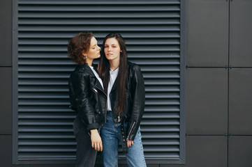 Fototapeta premium Portrait of two sexy girls in leather jackets, standing on a dark wall background and posing at camera. Pair of lesbians stands on black background. Pride concept. Beautiful girlfriend on background.