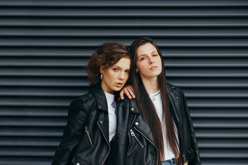 Fashionable portrait of two models with freckles in casual clothes on a dark street background, wearing leather jackets, looking into the camera with a serious face.Girl posing at camera on dark wall