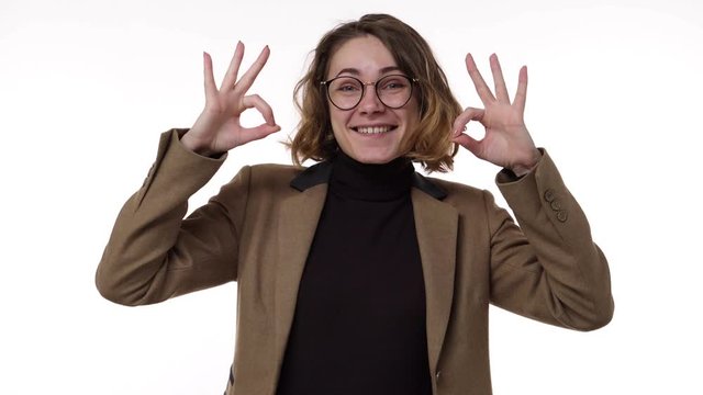 Beautiful young woman in brown jacket smiles and shows thumbs up gesture ank OK winking isolated on white background