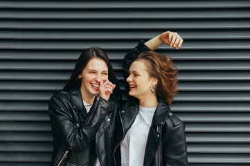 Happy girls in casual casual clothes, standing against a black wall background, talking and smiling, wearing leather jackets.Two cheerful girlfriends laughing on dark background. Copy space