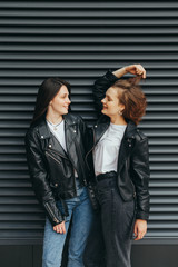 Happy couple girls in casual casual clothes are standing against a black wall background, talking and smiling, wearing leather jackets.Two cheerful girlfriends laughing on dark background. Vertical