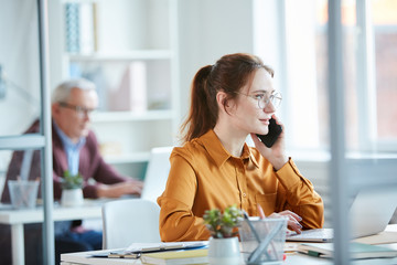 Young businesswoman sitting at her workplace in front of computer and talking on mobile phone at office