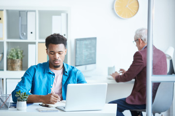 African young businessman working on laptop at the table with his colleague in the background