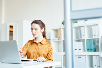 Serious young businesswoman sitting at the table in front of note pad looking at monitor of laptop and planning her work
