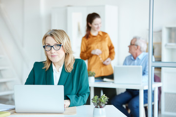 Portrait of mature businesswoman in eyeglasses sitting at the table in front of laptop and looking at camera with her colleagues in the background