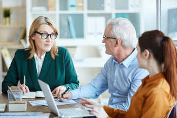 Obraz premium Mature businesswoman in eyeglasses talking to her colleagues and they discussing business plan at the table at business meeting