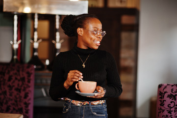 Obraz premium African woman in black sweater and eyeglasses posed at cafe with cup of hot drink.