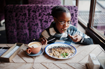 Fototapeta na wymiar African woman in checkered cape and eyeglasses sitting at cafe and eating dessert.