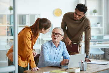 Obraz premium Young business people talking to their mature experienced colleague while he sitting at the table and using laptop
