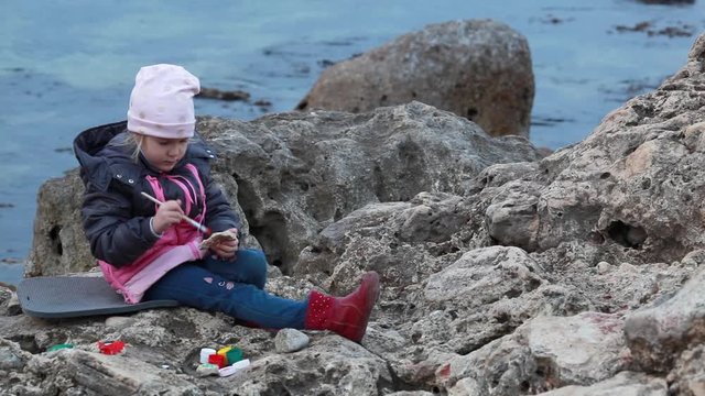 Girl paints pebbles on the seashore.