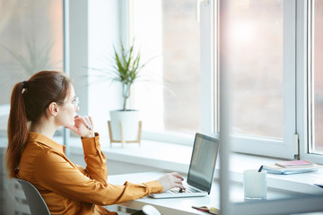 Young pensive businesswoman sitting at her workplace in front of the window and working on laptop computer at office