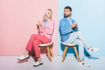 suspicious woman and handsome man holding smartphones on pink and blue background