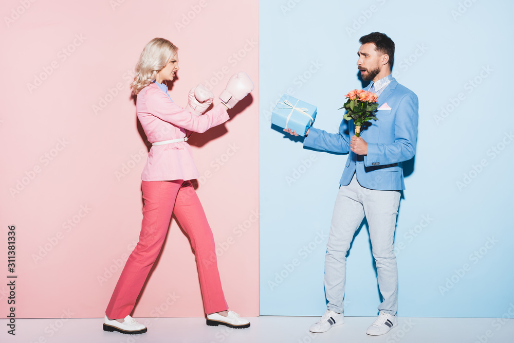 Wall mural angry woman with boxing gloves fighting with scared man with gift and bouquet on pink and blue backg