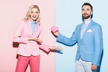 handsome man giving heart-shaped card to shocked woman on pink and blue background