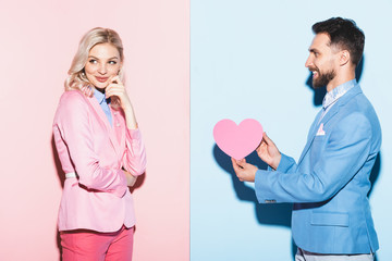 handsome man giving heart-shaped card to dreamy woman on pink and blue background