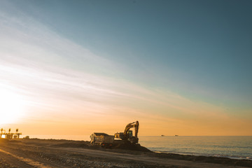 Fototapeta premium Excavator loads the excavation onto a truck (hydraulic)are heavy construction equipment consisting of an arrow,a bucket and a cabin on a rotating platform.On the beach with the sea and the setting sun