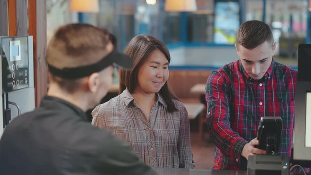 Young smiling positive couple pay for the order in cafe by credit debit card at counter after being served by a smiling barista in coffee shop fastfood cafe