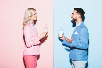 side view of attractive woman and smiling man holding cocktails on pink and blue background