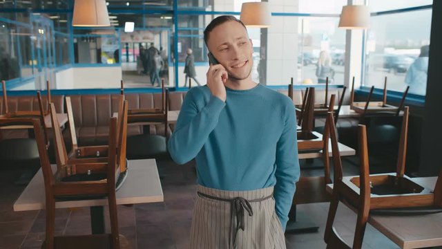Young caucasian waiter bartender or barista in uniform apron talking by mobile phone in empty cafe before opening. Owner of bar orders products from a supplier