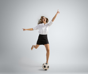 Born to be winner. Woman in office clothes kicking ball on grey studio background. Businesswoman training in motion, action. Unusual look for sport, new activity. Sport, football, healthy lifestyle.