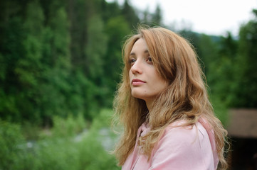 Naklejka premium Attractive young calm blonde woman in pink clothes with long hair on the hill with green forest background during spring or early autumn in the mountains.