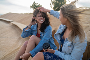Two beautiful girls blonde and brunette with a pretty smiles in denim jackets sitting on the wood bench talking laughing and the wind developing their hair
