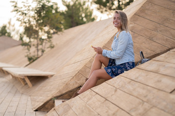 Beautiful smiley blonde girl in denim jacket and skirt sitting on the wood stairs, holding smartphone, looking at camera and the wind developing her hair. Student girl smiling.