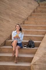 Beautiful blonde smiley girl in denim jacket and dress sitting on the wood stairs, holding smartphone, smiling and the wind developing her hair.