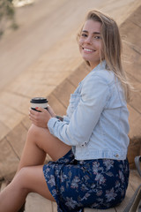 Close up portrait of beautiful blonde happy girl in a denim jacket and blue dress sitting on the stairs, looking at camera, fix her hair and drinking coffee from an eco cup.