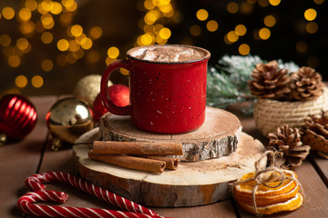Christmas drink. hot chocolalad with marshmallows in a beautiful red mug on a wooden table. nearby spruce cones, candy and dried oranges.
