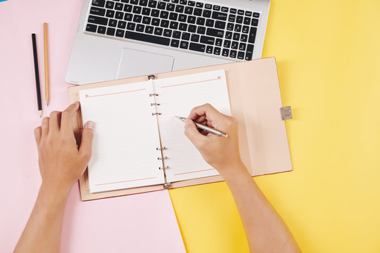 Hands of college student sitting at bright desk, working on laptop and writing in planner