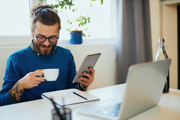 Young pensive attractive caucasian bearded freelancer sitting in his home office, using tablet for reading news and holding cup of coffee.
