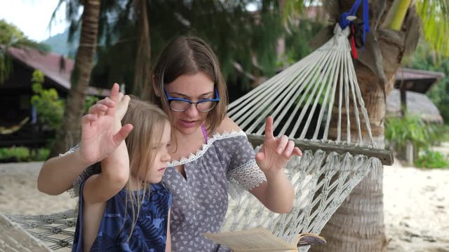 Mom Reads Book or Story Tale for Daughters on Sea Coast. Happy Family Sits On Hammock In Tropical Country. Nature and tropical palm trees background. Summer day outdoors