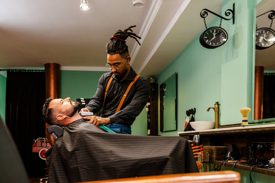 barbero afroamericano esta cortando y arreglando la barba a un hombre caucasico en una barber&iacute;a con una maquina de afeitar electrica. estilo hispter, moderno, vintage, retro.