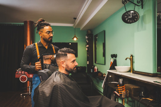 un barbero afroamericano de estilo hipster ense&ntilde;a a su cliente blanco caucasico con un espejo su corte de pelo y barba.