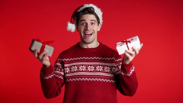 Young handsome man smiling and holding gift box on red studio background. Guy in Santa hat and ugly christmas sweater. New year mood.