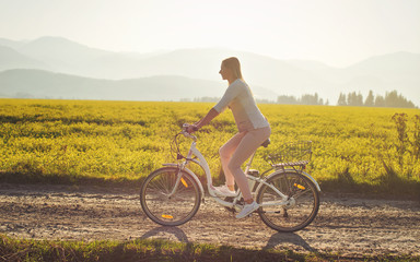 Obraz premium Young woman rides electric bike over dusty country road, strong afternoon sun backlight in background shines on yellow flowers field, view from side