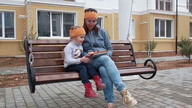 Mom and daughter sit on a swing and look at the tablet.