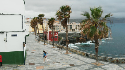 AERIAL. Cinematic shot - a girl walks through a deserted Spanish city, along the coast, during a strong wind, in overcast weather, a bird's-eye view. Siesta, Gran Canaria, Spain