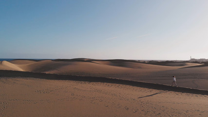 Aerial view is a side view of the dune along which a beautiful young woman walks. The golden hour at sunset or sunrise in the desert. Maspalomas, Gran Canaria
