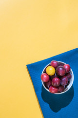 A many ripe dark blue plum stands in a white plate on a blue fabric. Fruit minimal concept on a yellow background. Vertical photo