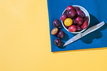 A many ripe dark blue plum stands in a white plate on a blue fabric. Plum is cut with a knife....