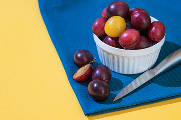 A many ripe dark blue plum stands in a white plate on a blue fabric. Plum is cut with a knife. Fruit minimal concept on a yellow background
