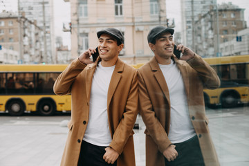 Cheerful happy positive young arabian man talking on phone. Stand outside on street and lean to glass wall. Reflection. Yellow bus passing behind. Big city life.