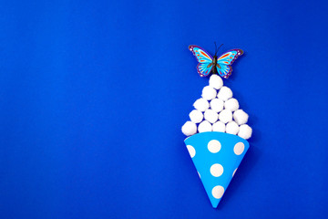 paper cone, blue in white peas,fluffy white balls, imitation of candy, ice cream, popcorn, butterfly sits on top,blue background