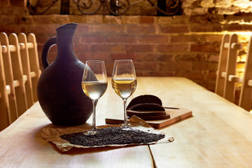 Two glasses of white wine, grape seeds, a clay jug and a few pieces of bread on a wooden table in a restaurant