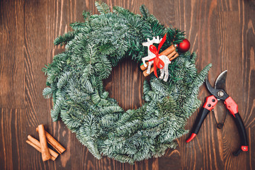 Top view florist making Christmas wreath on wooden table, flat lay tree branches secateurs, deer with red scarf