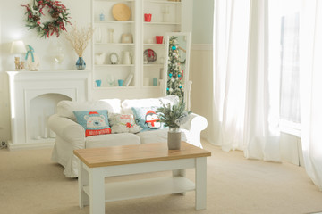 The interior of the Christmas room with a large window, a white sofa with pillows at the coffee table.