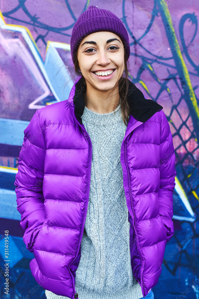 Wall mural outdoor portrait of young attractive woman wearing in violet down jacket and knitted hat, smiling to